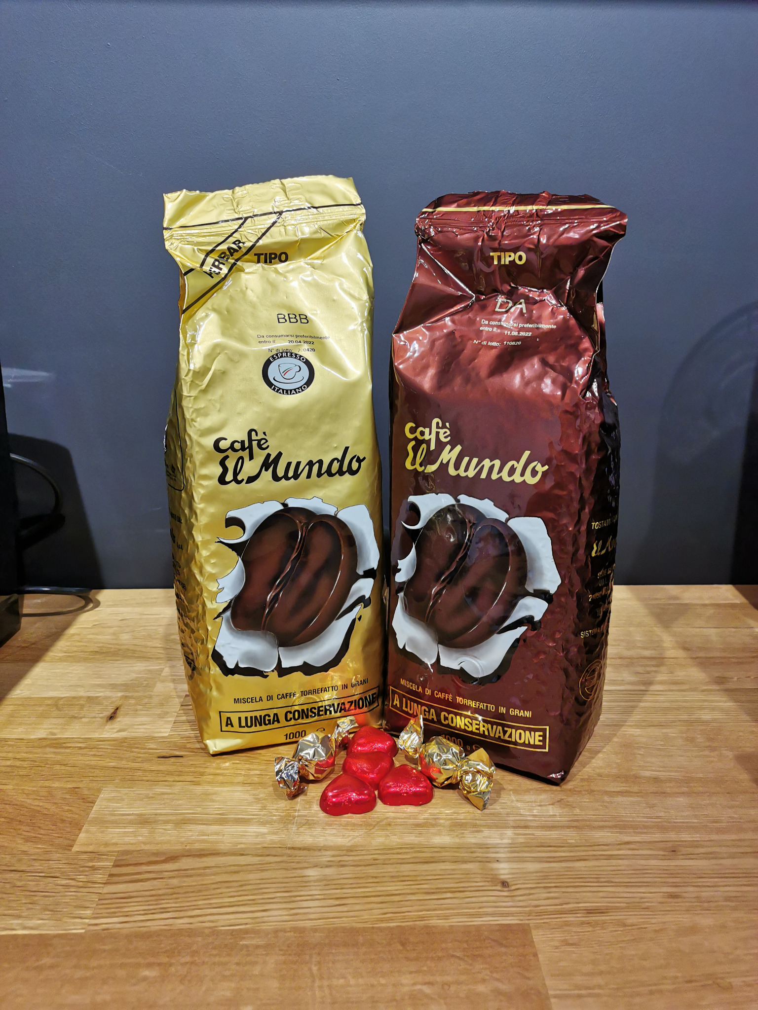 Dwa opakowania kawy ziarnistej Cafe El Mundo, jedno w złotym, drugie w bordowym opakowaniu, z czekoladkami w kształcie serc i złotych cukierków na drewnianym blacie.