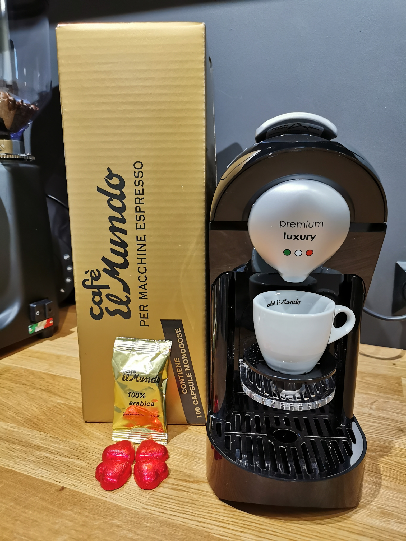 Czarny ekspres do kawy na kapsułki z filiżanką Cafe El Mundo, obok złote pudełko i saszetka kawy oraz dwie czerwone czekoladki w kształcie serca.