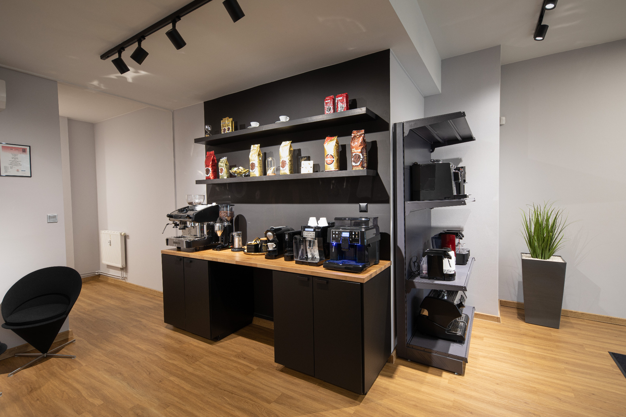 Wnętrze biura lub sklepu z ekspresami do kawy, prezentacja różnych modeli ekspresów na blacie i półkach, obok widoczny fotel i doniczka z rośliną.