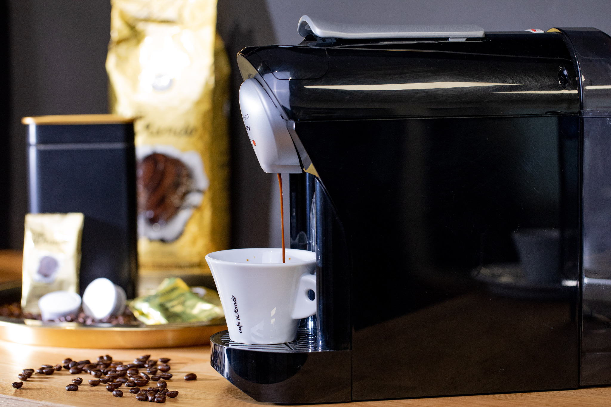 Czarny ekspres automatyczny nalewa kawę do białej filiżanki z napisem 'Cafe el Mundo', w tle złote opakowanie kawy i czarna puszka na tacy, rozsypane ziarna kawy na drewnianym blacie.