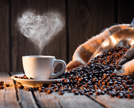 Biała filiżanka z parującą kawą, której para układa się w kształt serca, obok jutowy worek z ziarnami kawy i metalowa łopatka na drewnianym stole.