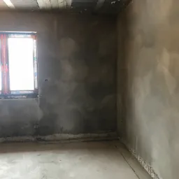 Surowe wnętrze w trakcie prac wykończeniowych; ściany pokryte świeżym, szarym tynkiem, widoczne kable elektryczne przy podłodze, okno z naklejkami producenta.