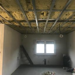 Tynki Maszynowe - Surowy stan wnętrza z widoczną konstrukcją sufitu podwieszanego z izolacją z wełny mineralnej, nieotynkowane ściany, okno z żaluzjami, drabina oparta o ścianę, instalacja elektryczna w suficie...