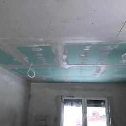 Tynki Maszynowe - Sufit z płyt gipsowo-kartonowych w trakcie montażu, widoczne łączenia wzmocnione taśmą i szpachlą, zwisające kable elektryczne, surowe ściany przed malowaniem, okno z roletami w tle.