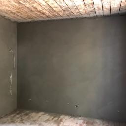 Tynki Maszynowe - Surowe wnętrze pokoju po nałożeniu szarego tynku, widoczne fragmenty cegieł i drewniany sufit, instalacja elektryczna wystająca ze ściany.