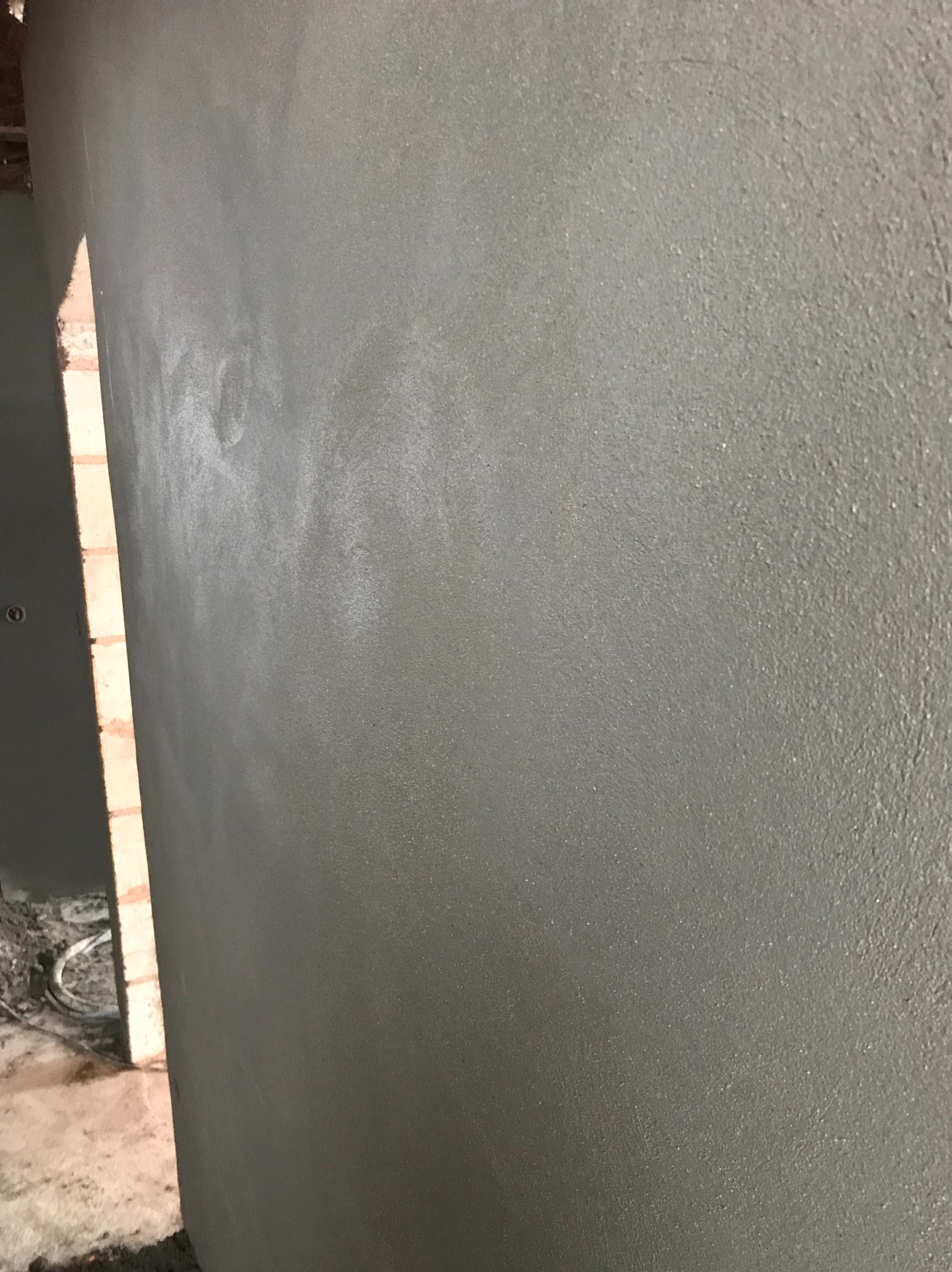 Świeżo nałożony, szary tynk na ścianie z widoczną strukturą, sąsiadujący z fragmentem muru z czerwonej cegły i elementami instalacji elektrycznej w tle.
