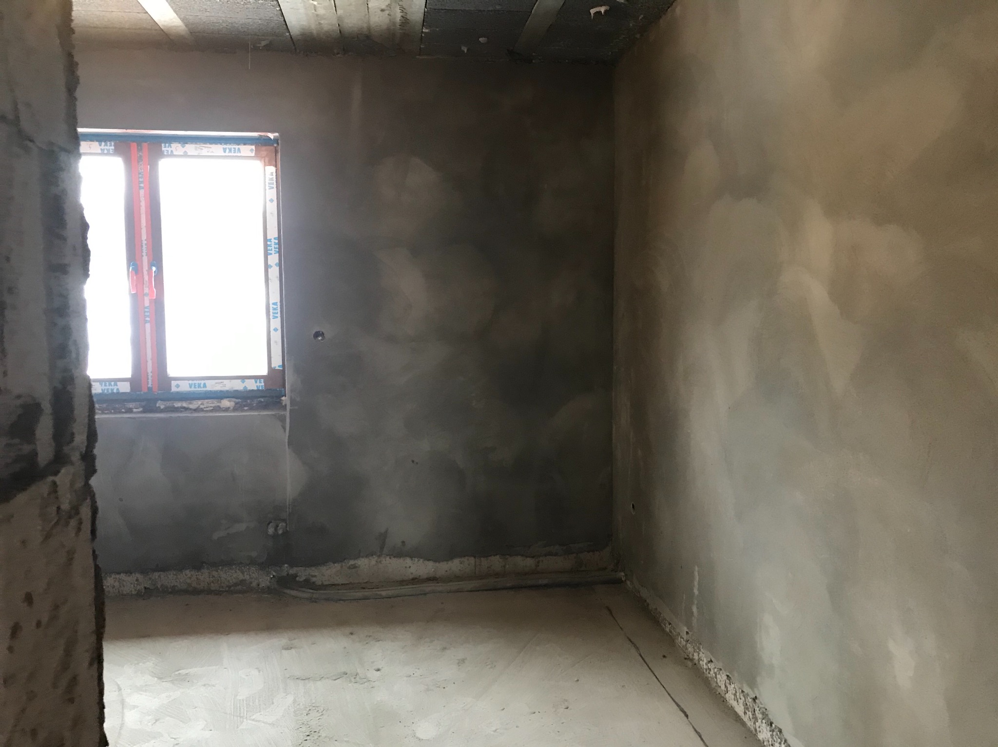 Surowe wnętrze w trakcie prac wykończeniowych; ściany pokryte świeżym, szarym tynkiem, widoczne kable elektryczne przy podłodze, okno z naklejkami producenta.