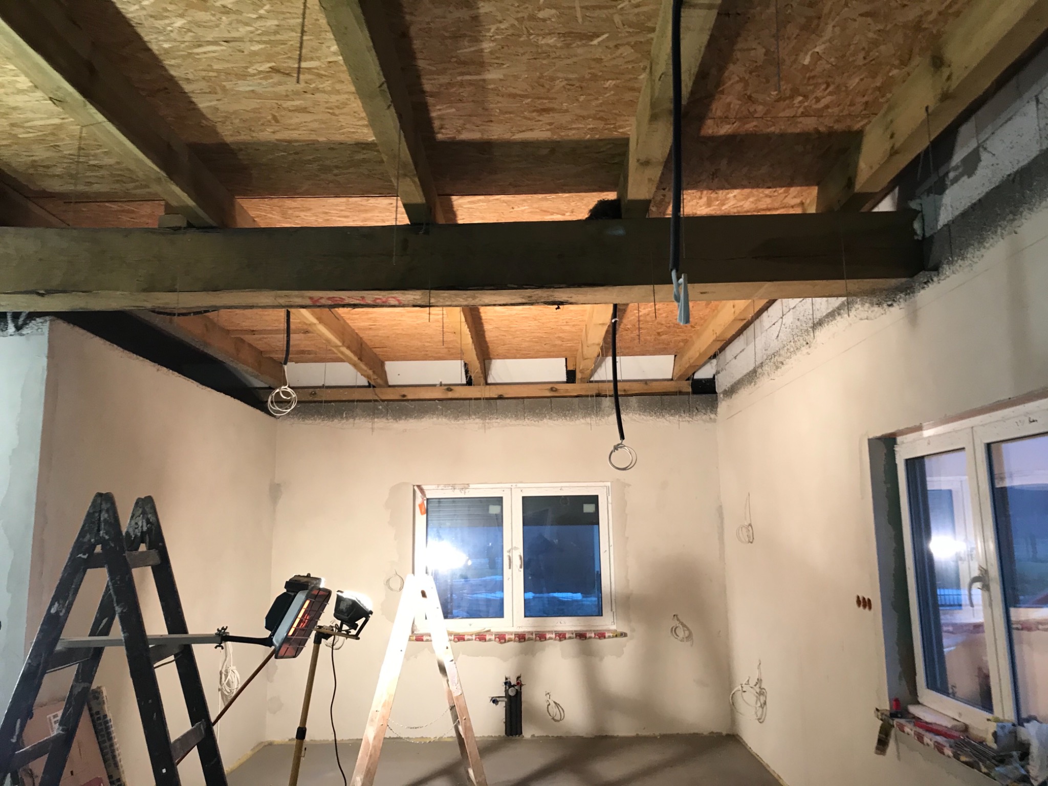 Wnętrze w trakcie wykańczania: surowy strop drewniany, ściany po tynkowaniu, widoczne instalacje elektryczne, drabiny i lampa budowlana.