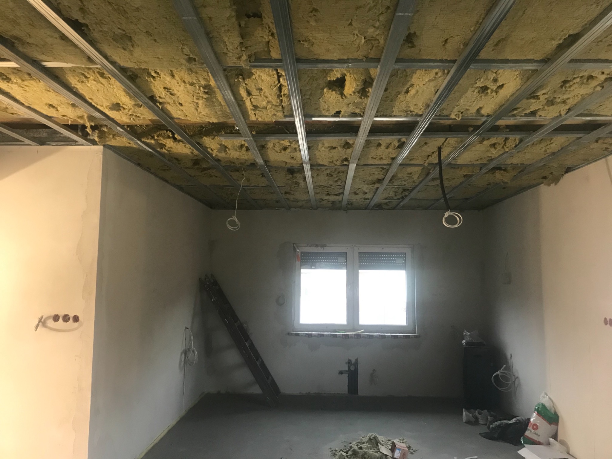 Surowy stan wnętrza z widoczną konstrukcją sufitu podwieszanego z izolacją z wełny mineralnej, nieotynkowane ściany, okno z żaluzjami, drabina oparta o ścianę, instalacja elektryczna w suficie...