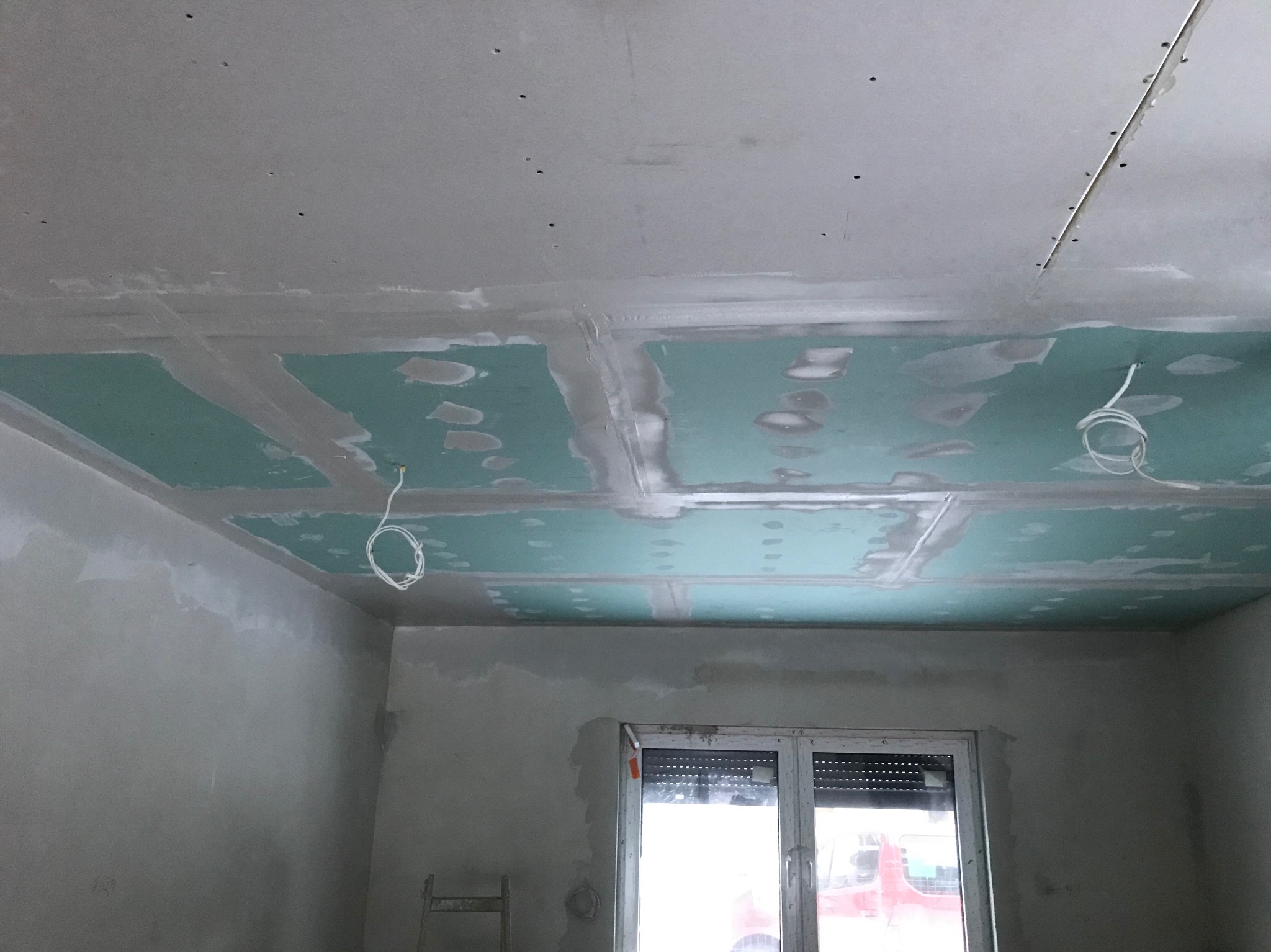 Sufit z płyt gipsowo-kartonowych w trakcie montażu, widoczne łączenia wzmocnione taśmą i szpachlą, zwisające kable elektryczne, surowe ściany przed malowaniem, okno z roletami w tle.