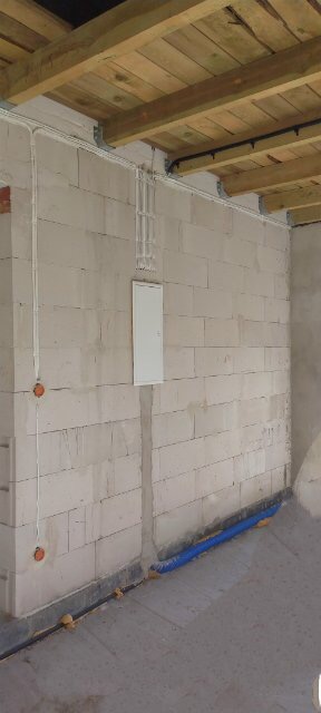 Surowa ściana z betonu komórkowego z widoczną instalacją elektryczną w peszlach, skrzynką rozdzielczą i puszkami pod gniazdka, pod drewnianym stropem.