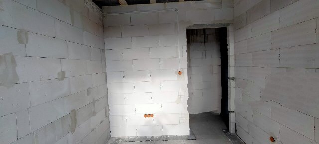 Surowe wnętrze z białymi ścianami z betonu komórkowego, widoczne otwory pod instalacje elektryczne i przygotowane nadproże drzwiowe.