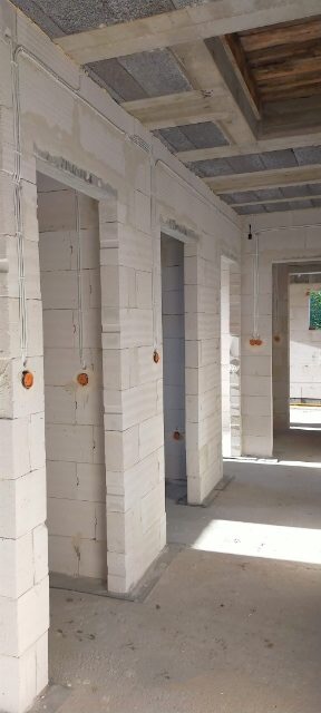 Wnętrze w stanie surowym: ściany z białego betonu komórkowego, widoczne otwory drzwiowe, instalacja elektryczna w peszlach, sufit betonowy z widocznymi elementami drewnianymi.