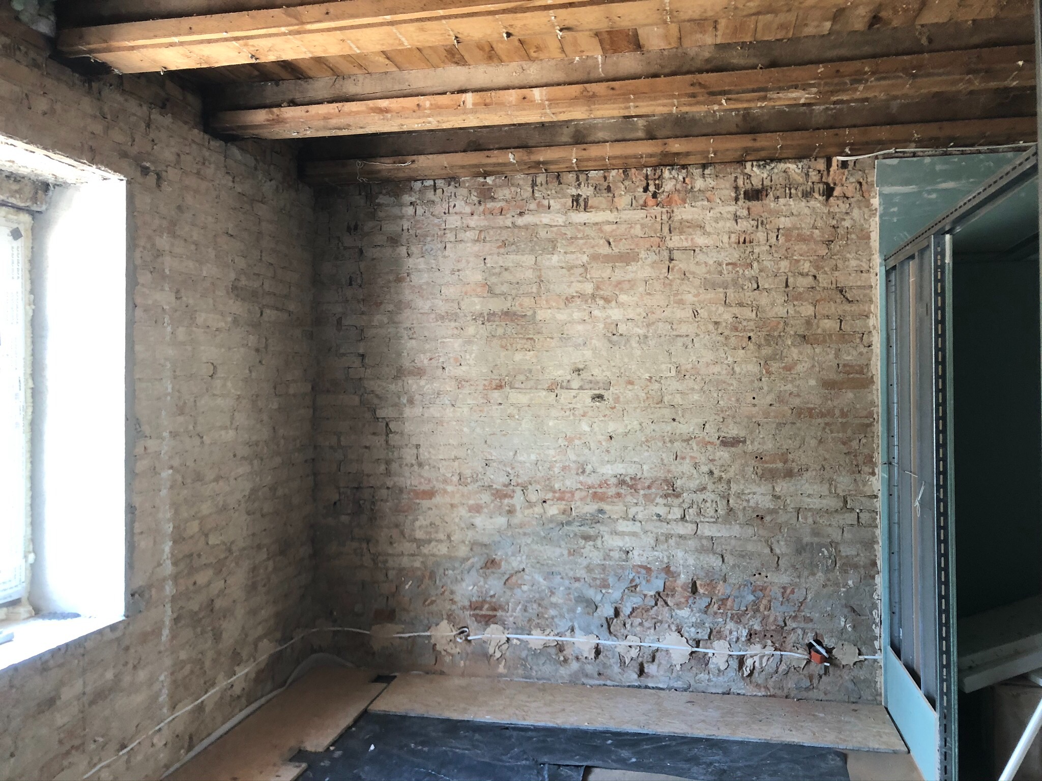 Surowe wnętrze z odsłoniętą cegłą, drewnianym stropem i przygotowanym podłożem pod podłogę, widoczna instalacja elektryczna.