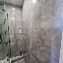 Szara łazienka z kabiną prysznicową typu walk-in, wykończona płytkami imitującymi beton, z widocznymi włącznikami światła i minimalistyczną armaturą.