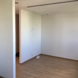 Pomalowane na biało wnętrze pokoju z drewnianą podłogą, widoczne gniazdka elektryczne i ślady po wierceniu, z resztkami materiałów budowlanych przy ścianie.