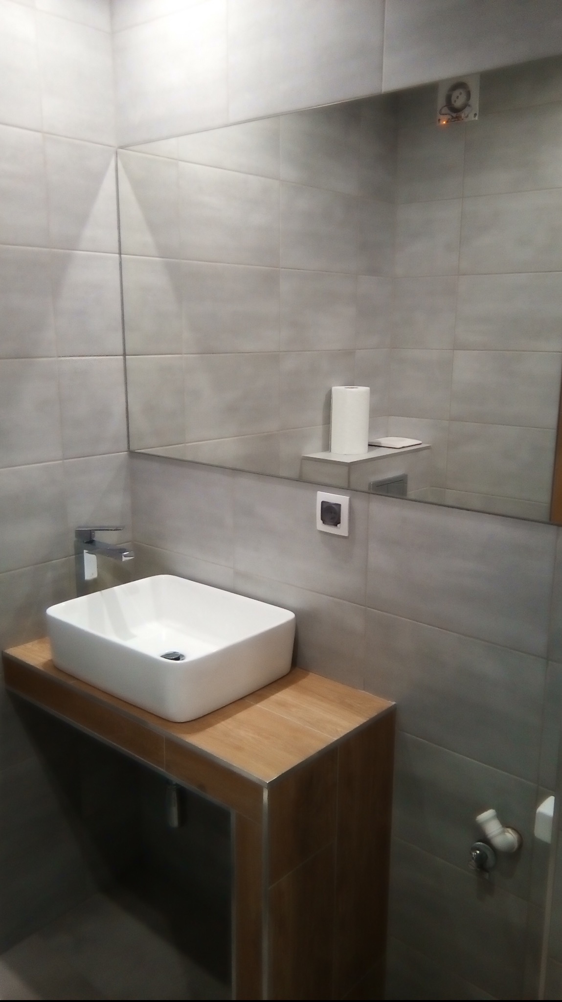 Nowoczesna łazienka z kwadratową umywalką nablatową, drewnianym blatem i dużym lustrem na szarych płytkach, widoczne detale instalacji.