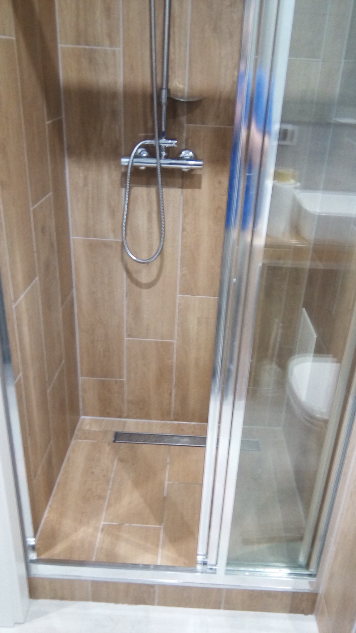 Wykończona kabina prysznicowa z brązowymi płytkami imitującymi drewno, chromowaną armaturą i odpływem liniowym w podłodze. Widoczna szklana ściana kabiny z aluminiową ramą.