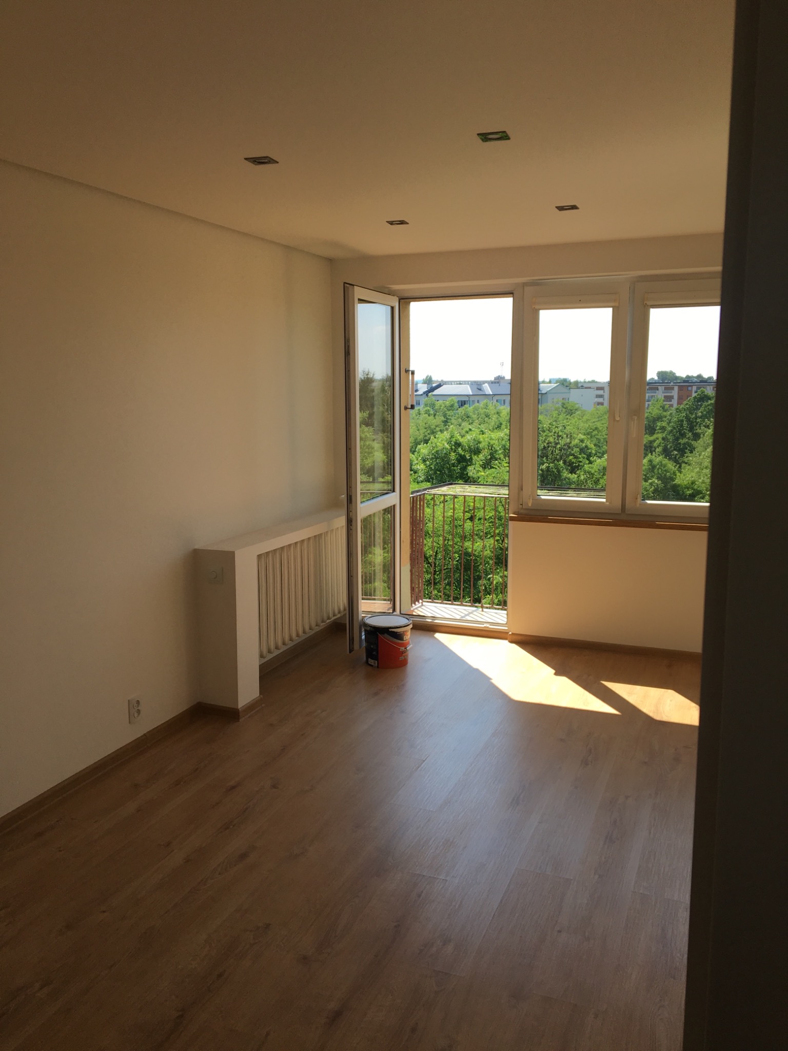 Jasne, puste pomieszczenie z drewnianą podłogą i otwartym balkonem; wiadro z farbą stoi przy drzwiach balkonowych, ściany pomalowane na jasny kolor, wpadające promienie słońca.