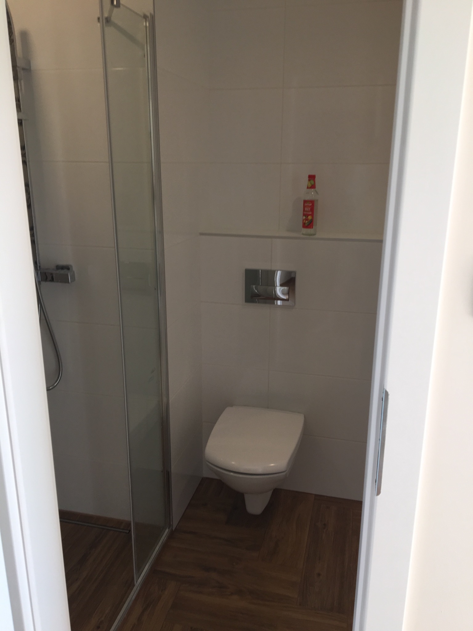 Nowoczesna łazienka z kabiną prysznicową i podwieszaną toaletą, wykończona białymi płytkami i drewnopodobną podłogą, widok zza otwartych drzwi, butelka płynu na półce.