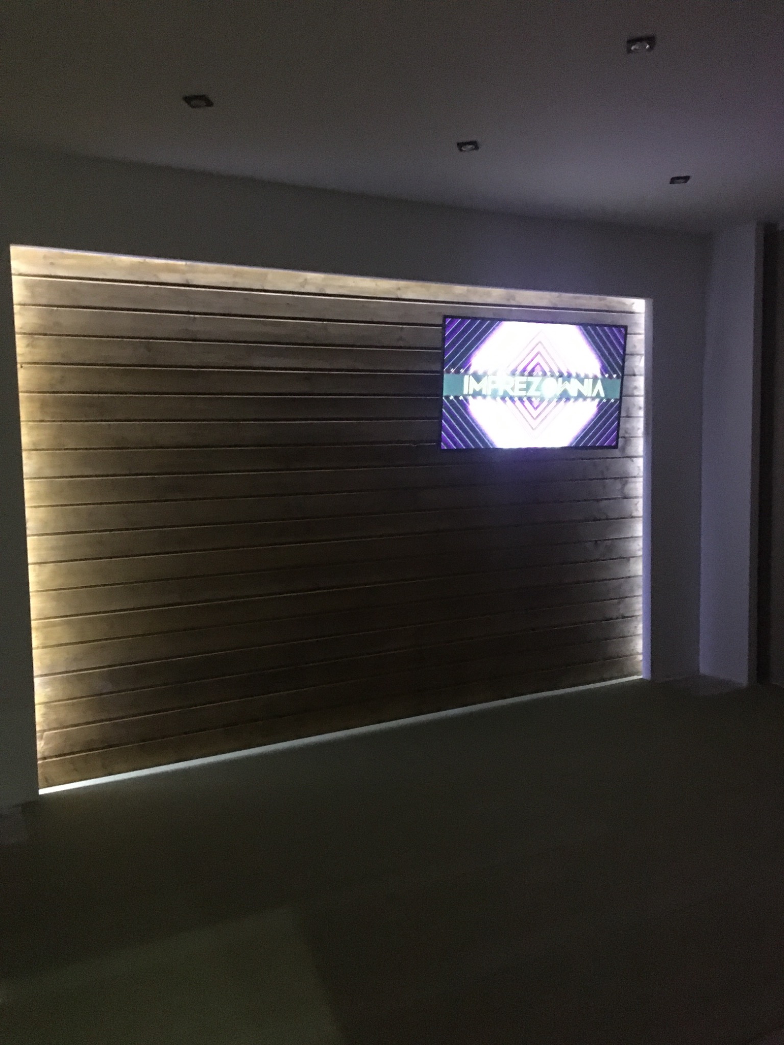 Ściana z drewnianych paneli z podświetleniem LED po bokach, z wbudowanym telewizorem wyświetlającym napis 'IMPREZOWNIA' w neonowych kolorach, oświetlenie sufitowe punktowe