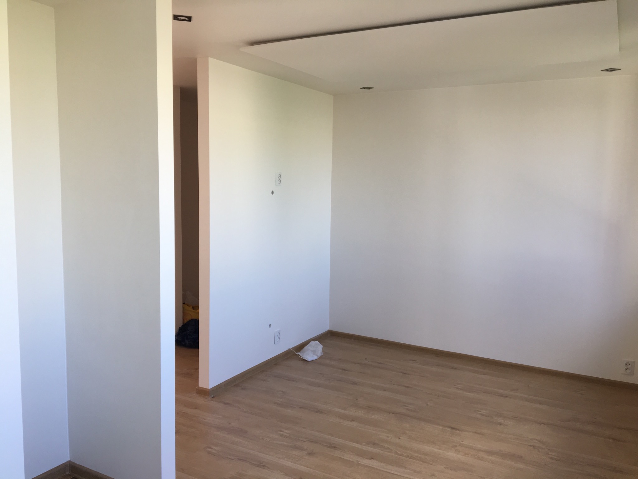 Pomalowane na biało wnętrze pokoju z drewnianą podłogą, widoczne gniazdka elektryczne i ślady po wierceniu, z resztkami materiałów budowlanych przy ścianie.