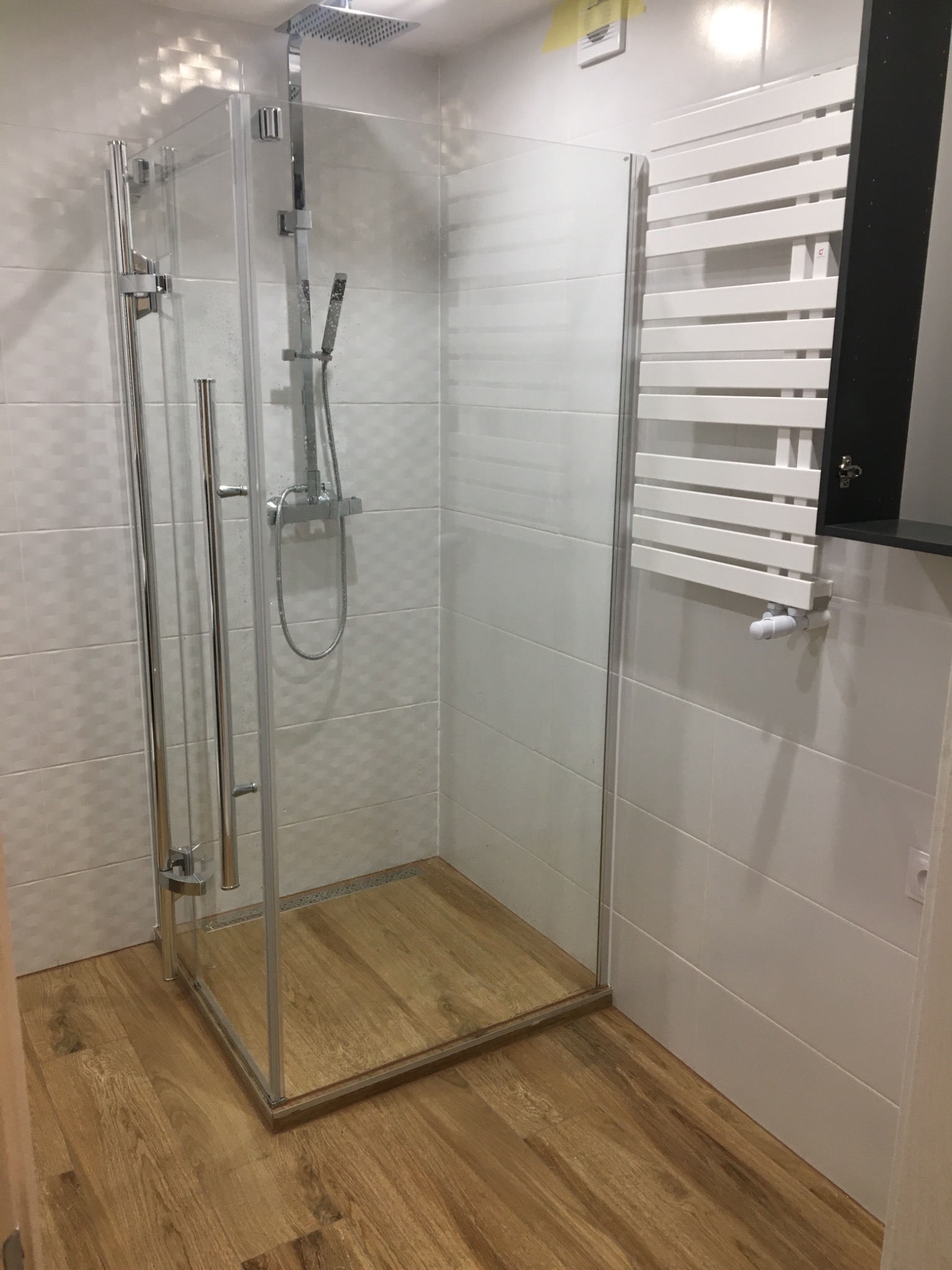 Nowoczesna kabina prysznicowa typu walk-in z drewnianym brodzikiem, białe płytki na ścianach i grzejnik drabinkowy w łazience.