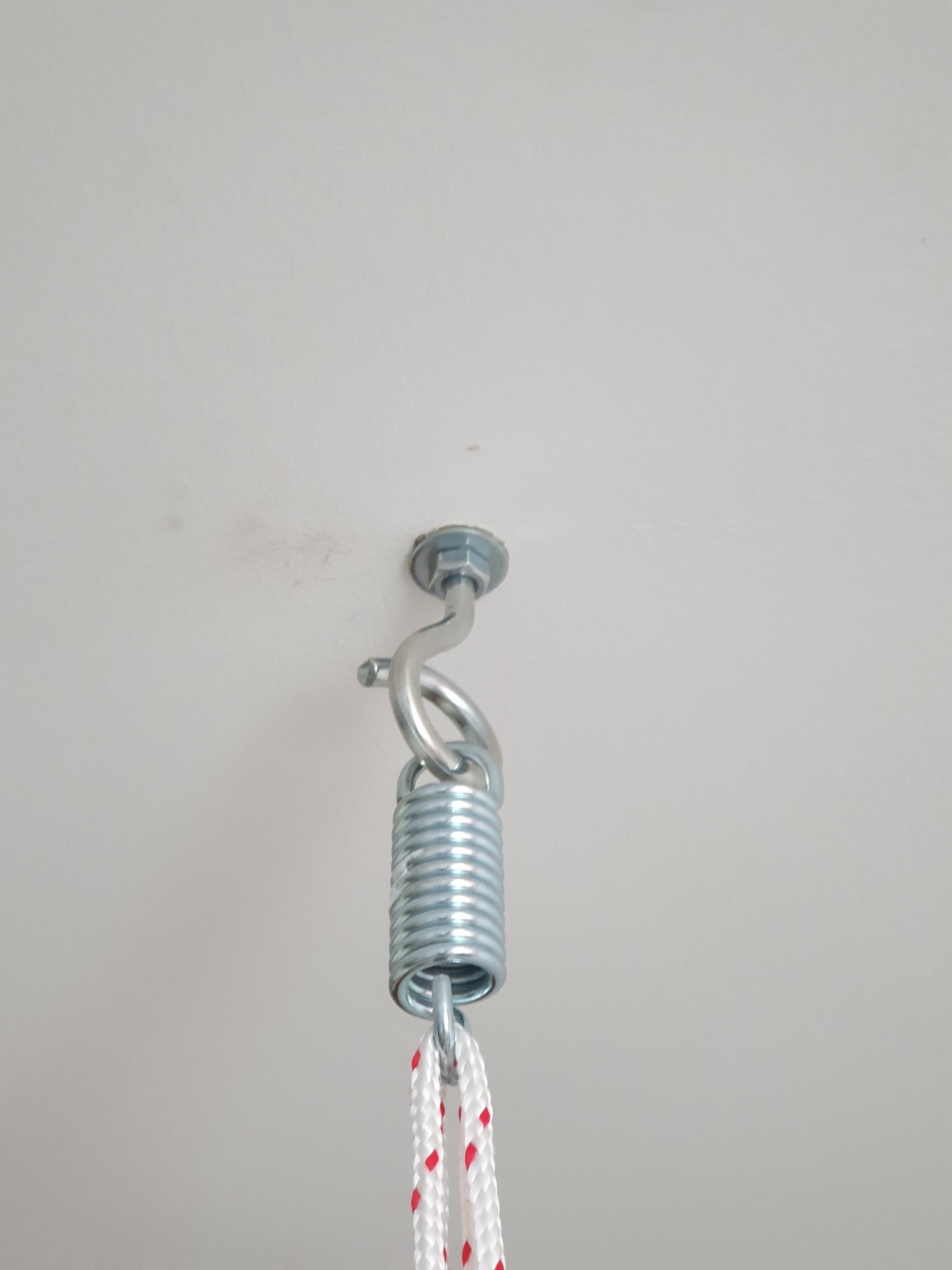 Metalowy hak z kołkiem rozporowym przymocowany do sufitu, połączony ze sprężyną i białą liną z czerwonymi akcentami, gotowy do zawieszenia huśtawki lub hamaka.