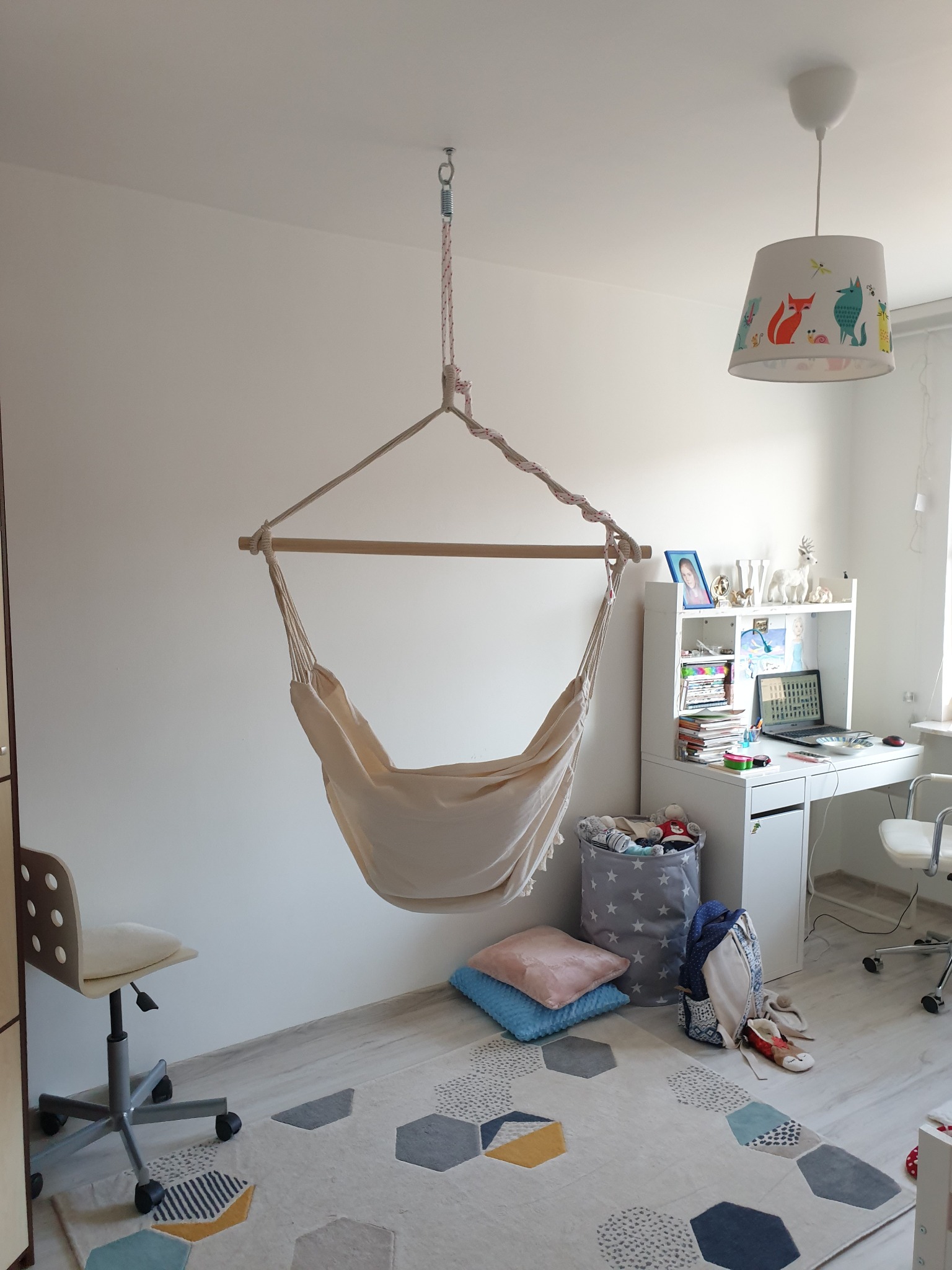 Beżowy hamak fotel wiszący na linie w pokoju dziecięcym, pod nim poduszki i kosz na zabawki, obok biurko z laptopem, całość na dywanie w geometryczne wzory.