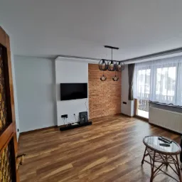 Salon po remoncie: ściana z cegły dekoracyjnej, gładkie białe ściany, drewniana podłoga, obniżony sufit z oświetleniem LED, czarny telewizor na ścianie, nowoczesna lampa wisząca z okrągłymi...