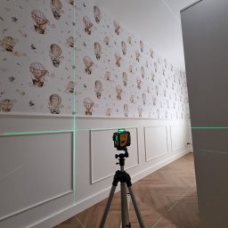 Perfekt Mal Adrian Szpora - Ściana z tapetą w balony i sztukaterią, oświetlona laserem krzyżowym na statywie, wskazującym poziom. Parkiet w jodełkę na podłodze. Widok z perspektywy korytarza.