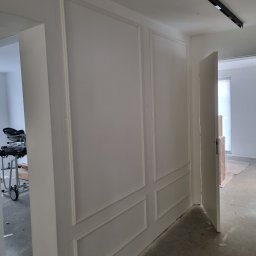 Perfekt Mal Adrian Szpora - Biała ściana z dekoracyjnymi panelami w stylu klasycznym, widoczna w trakcie wykańczania wnętrza. Otwarta przestrzeń z widocznym sprzętem budowlanym w tle.