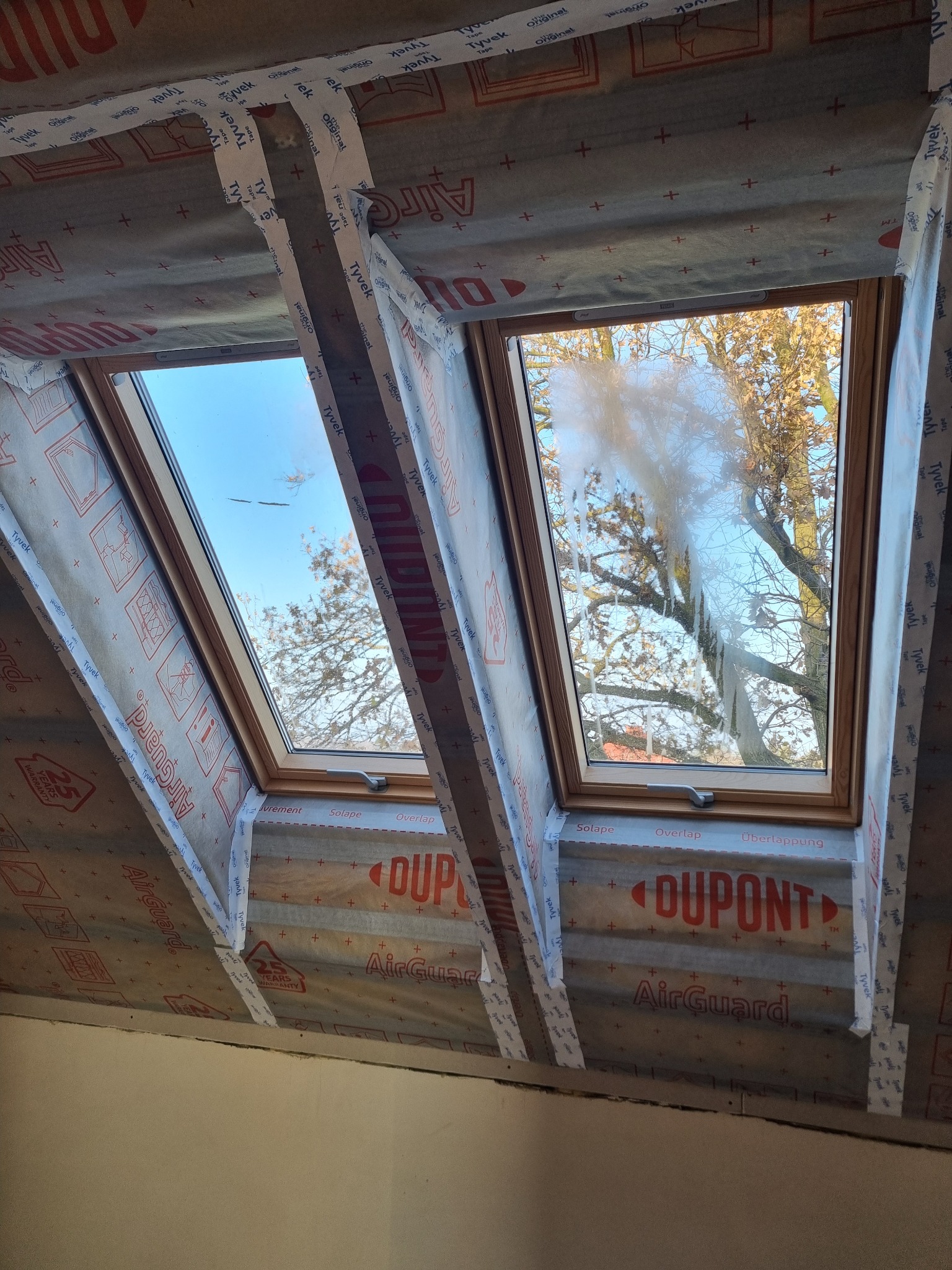 Dwa okna dachowe w trakcie montażu, zabezpieczone folią i taśmą Tyvek, widok od wewnątrz na niebo i drzewa. Etap prac wykończeniowych, izolacja poddasza.
