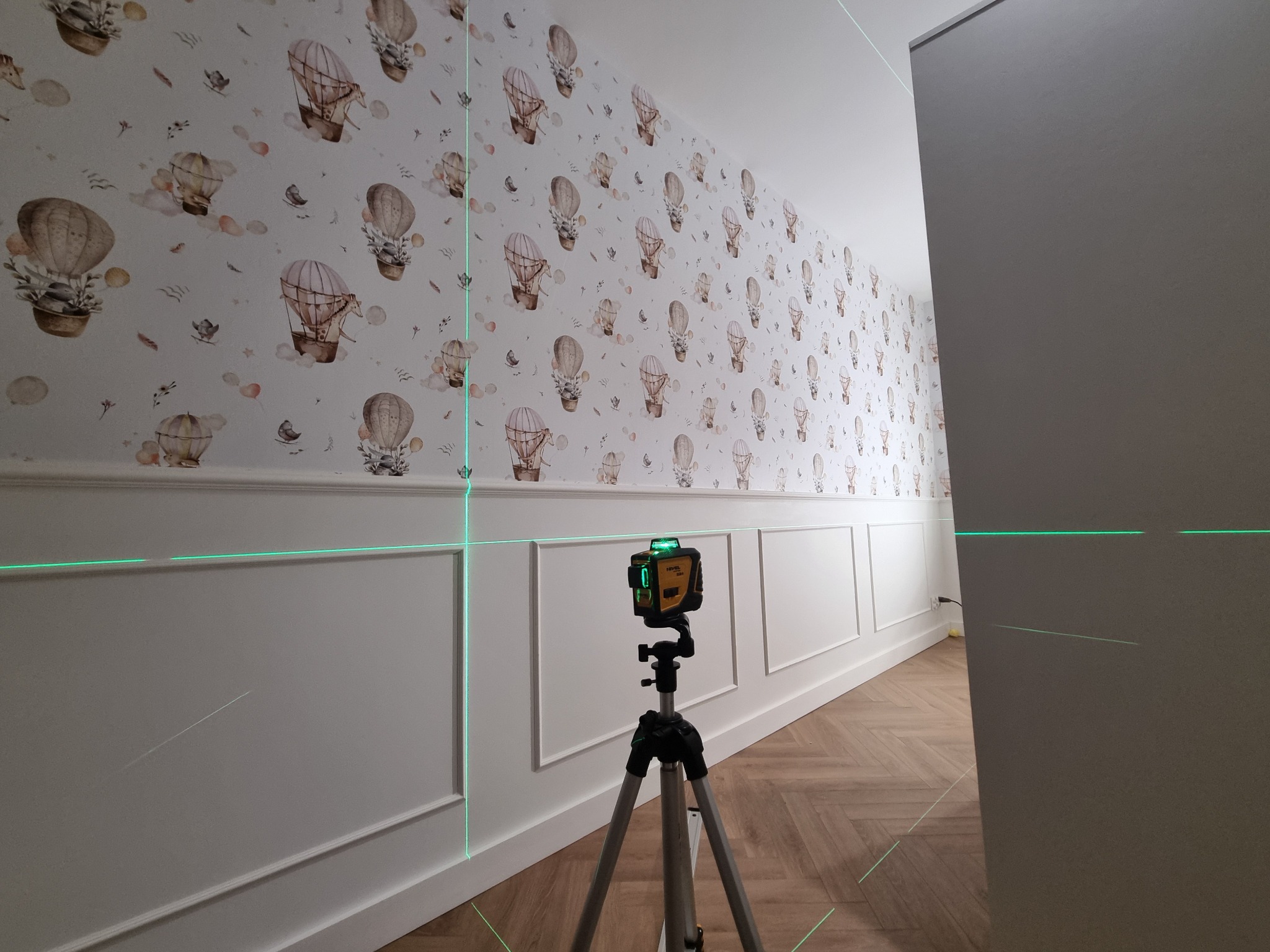 Ściana z tapetą w balony i sztukaterią, oświetlona laserem krzyżowym na statywie, wskazującym poziom. Parkiet w jodełkę na podłodze. Widok z perspektywy korytarza.