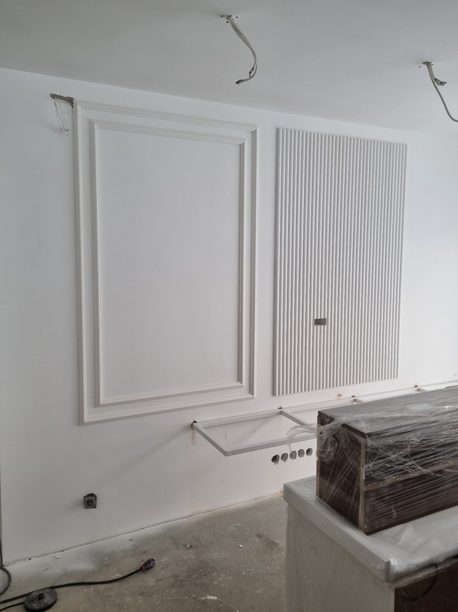 Białe panele dekoracyjne na ścianie w trakcie montażu, widoczne elementy instalacji elektrycznej i fragmenty mebli zabezpieczonych folią. Minimalistyczny design.