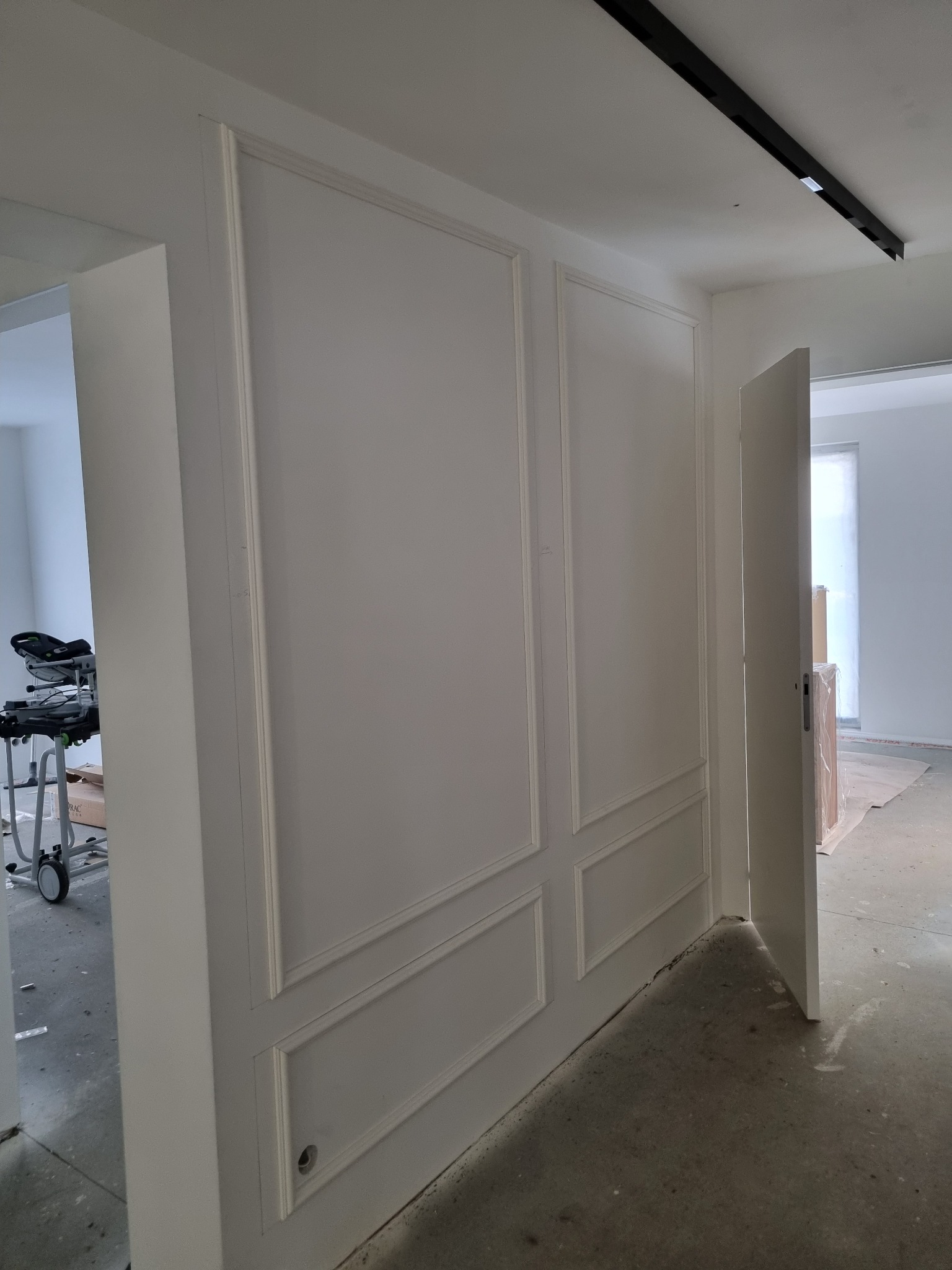 Biała ściana z dekoracyjnymi panelami w stylu klasycznym, widoczna w trakcie wykańczania wnętrza. Otwarta przestrzeń z widocznym sprzętem budowlanym w tle.