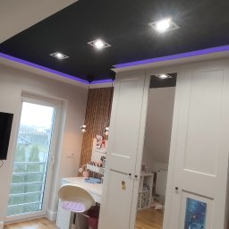 FachGro usługi remontowe - Pokój na poddaszu z czarnym sufitem, oświetleniem LED i drewnianą ścianą. Widoczne biurko, szafa z lustrem i telewizor. Nowoczesny design z elementami dekoracyjnymi.