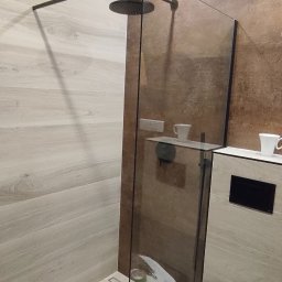 FachGro usługi remontowe - Nowoczesna kabina prysznicowa typu walk-in z czarną armaturą i szklaną ścianką. Na sedesie leżą narzędzia i akcesoria montażowe. Widoczne płytki imitujące drewno.