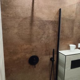 FachGro usługi remontowe - Nowoczesna kabina prysznicowa walk-in z brązowymi płytkami imitującymi kamień, czarną armaturą i szklaną ścianką. Widoczny odpływ liniowy i fragment szafki umywalkowej.