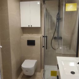 FachGro usługi remontowe - Nowoczesna łazienka z beżowymi płytkami, kabiną prysznicową z czarnymi ramami, podwieszaną toaletą i białą szafką z umywalką. Minimalistyczne oświetlenie.