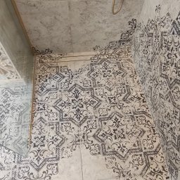 FachGro usługi remontowe - Wykończona kabina prysznicowa z dekoracyjnymi płytkami w odcieniach szarości i beżu, złota armatura. Widoczne stopy osoby robiącej zdjęcie.