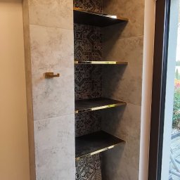 FachGro usługi remontowe - Narożna półka w łazience wyłożonej płytkami imitującymi kamień, z czarnym wnętrzem i złotymi krawędziami. Widoczne akcesoria łazienkowe i fragment okna.