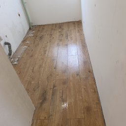 FachGro usługi remontowe - Układanie podłogi z paneli imitujących drewno w trakcie prac wykończeniowych. Widoczne białe krzyżyki dystansowe i surowe ściany w pomieszczeniu.