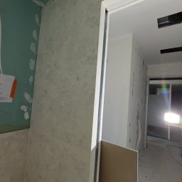 FachGro usługi remontowe - Ściana z płyt gipsowo-kartonowych w trakcie wykończenia, widoczne łączenia i szpachlowanie. W tle ościeżnica drzwiowa i fragment instalacji elektrycznej.