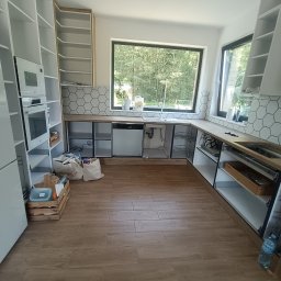 FachGro usługi remontowe - Wnętrze kuchni w trakcie montażu: białe szafki, drewniany blat, heksagonalne płytki na ścianie. Widoczne okno z widokiem na zieleń i drewniana podłoga.