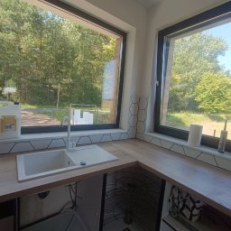 FachGro usługi remontowe - Jasna kuchnia z drewnianym blatem, białym zlewem i heksagonalnymi płytkami pod oknem z widokiem na zieleń. Kanister z płynem Axton na blacie.