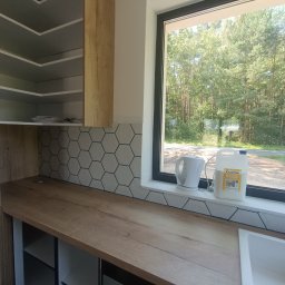 FachGro usługi remontowe - Jasna kuchnia z heksagonalnymi płytkami nad blatem, drewniane szafki i blat. Za oknem widok na zieleń. Kanister z preparatem do czyszczenia na parapecie.