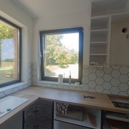 FachGro usługi remontowe - Jasna kuchnia z drewnianym blatem i heksagonalnymi płytkami. Widok na zieleń za oknem. Umywalka z białą baterią. Częściowo zamontowane szafki wiszące.