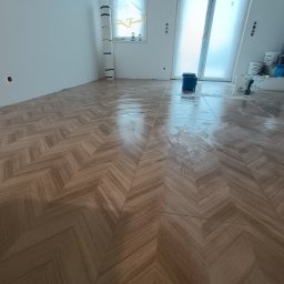 FachGro usługi remontowe - Ukończona podłoga z płytek ułożonych w jodełkę, z widocznymi akcesoriami malarskimi i zabezpieczonym oknem folią malarską. Jasne wnętrze w trakcie wykańczania.