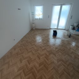 FachGro usługi remontowe - Ukończona podłoga z parkietu ułożonego w jodełkę w jasnym pokoju z zabezpieczonymi oknami i drzwiami. Widoczne narzędzia malarskie.